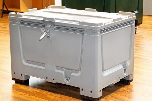 A large, light grey plastic skip with a rectangular shape, situated indoors on a polished wooden floor. The skip features reinforced edges and a secure, lid with handles on each side, designed for waste collection or disposal. The surface of the skip has a slightly textured finish, and the lid is closed, with a metal latch visible on the front side. The background includes a section of a green waste container and some other indistinct objects, suggesting a common storage or waste management area. The skip appears clean and well-maintained, suitable for commercial waste collection services, potentially hire or disposal in a UK town like Wallington, as part of services offered by Commercial Waste Wallington.