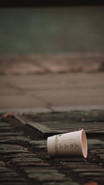 A discarded paper coffee cup lies tipped over on a paved outdoor surface, positioned on its side with the opening facing away from the camera. The cup appears to be made of disposable material with a 